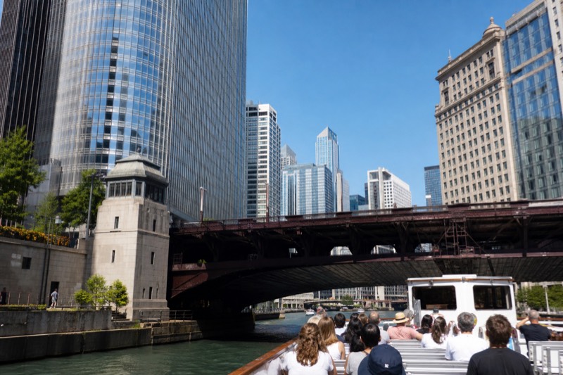 A tour boat passing beneath a low steel drawbridge on the Chicago River under bright midday sun, high-rises framing the view